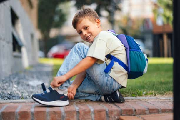 Young boy sits smiling outside, adjusting his shoes with a backpack, ready for a new school year. A joyful moment capturing the excitement of back-to-school preparations.